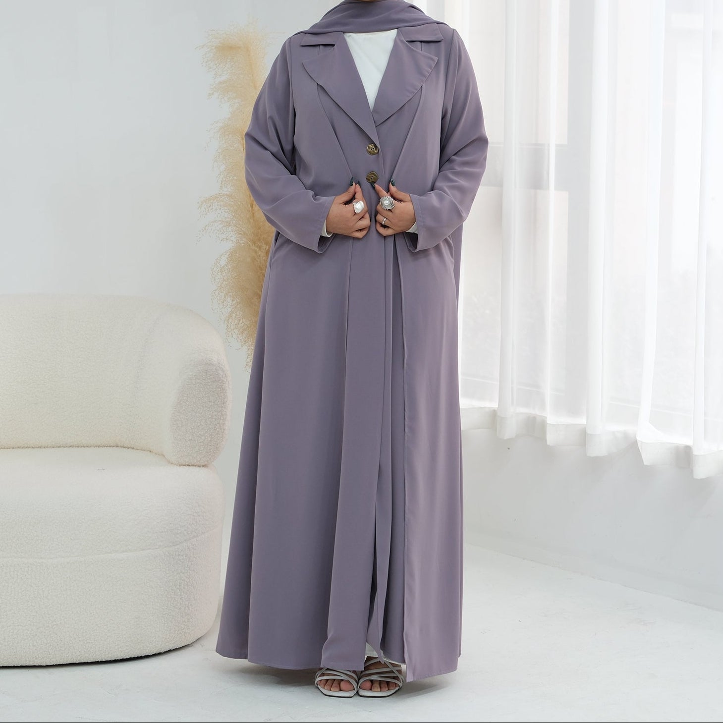 Woman wearing a long, light purple coat in a bright room with white curtains.