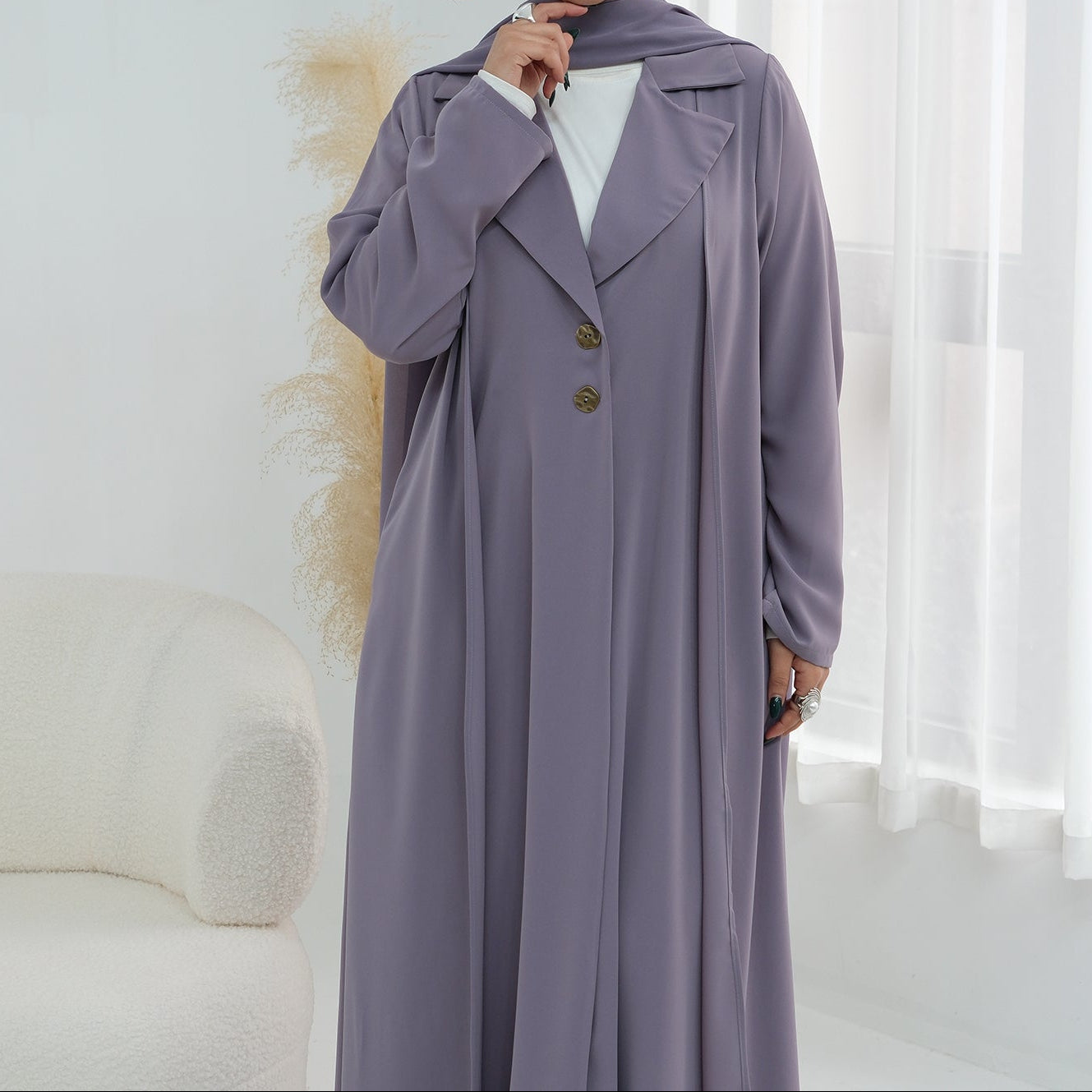 Woman wearing a long, light purple coat with a white shirt underneath, standing in a room with a white curtain.