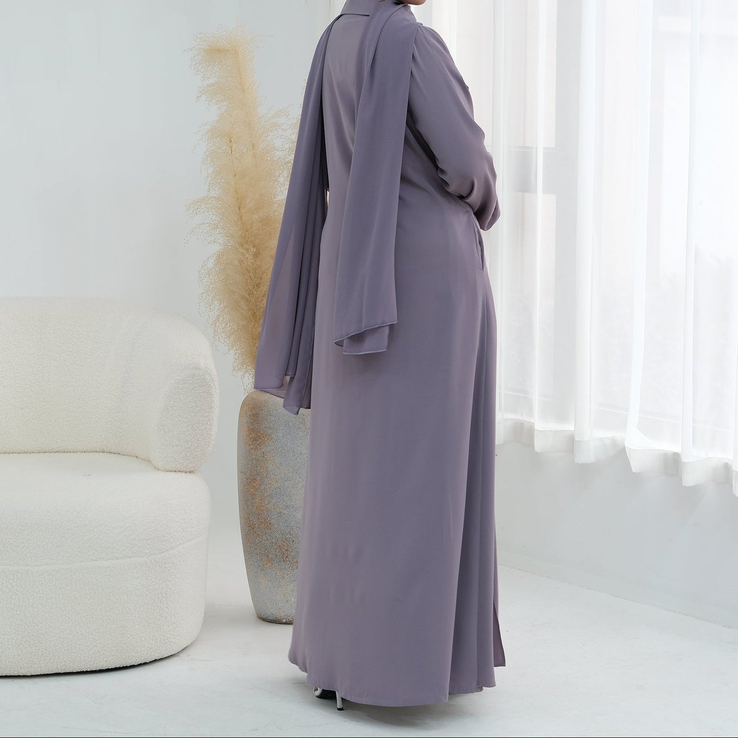 Woman in a long, light purple dress standing in a minimalistic room with white furniture and decor.