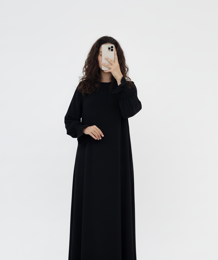 Person wearing a black abaya standing next to a vase with dried plants on a white background