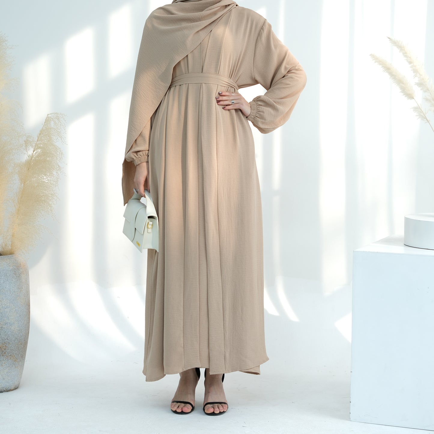 Person wearing a beige long dress with a matching hijab in a minimalistic setting. Noor Al Abaya (نور للعباية) - Noor & Grace