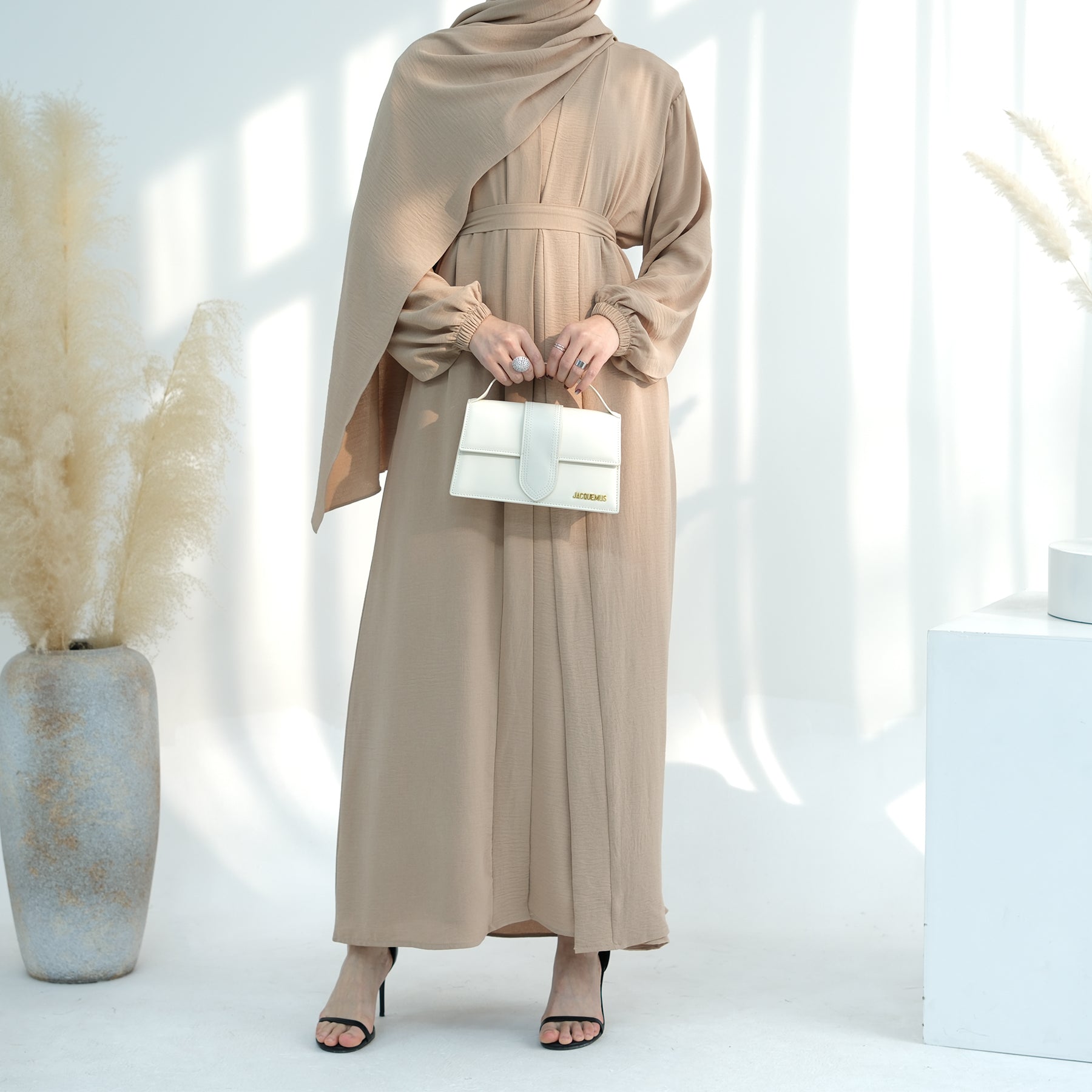 Woman in a beige abaya holding a white handbag in a minimalistic setting. Noor Al Abaya (نور للعباية) - Noor & Grace