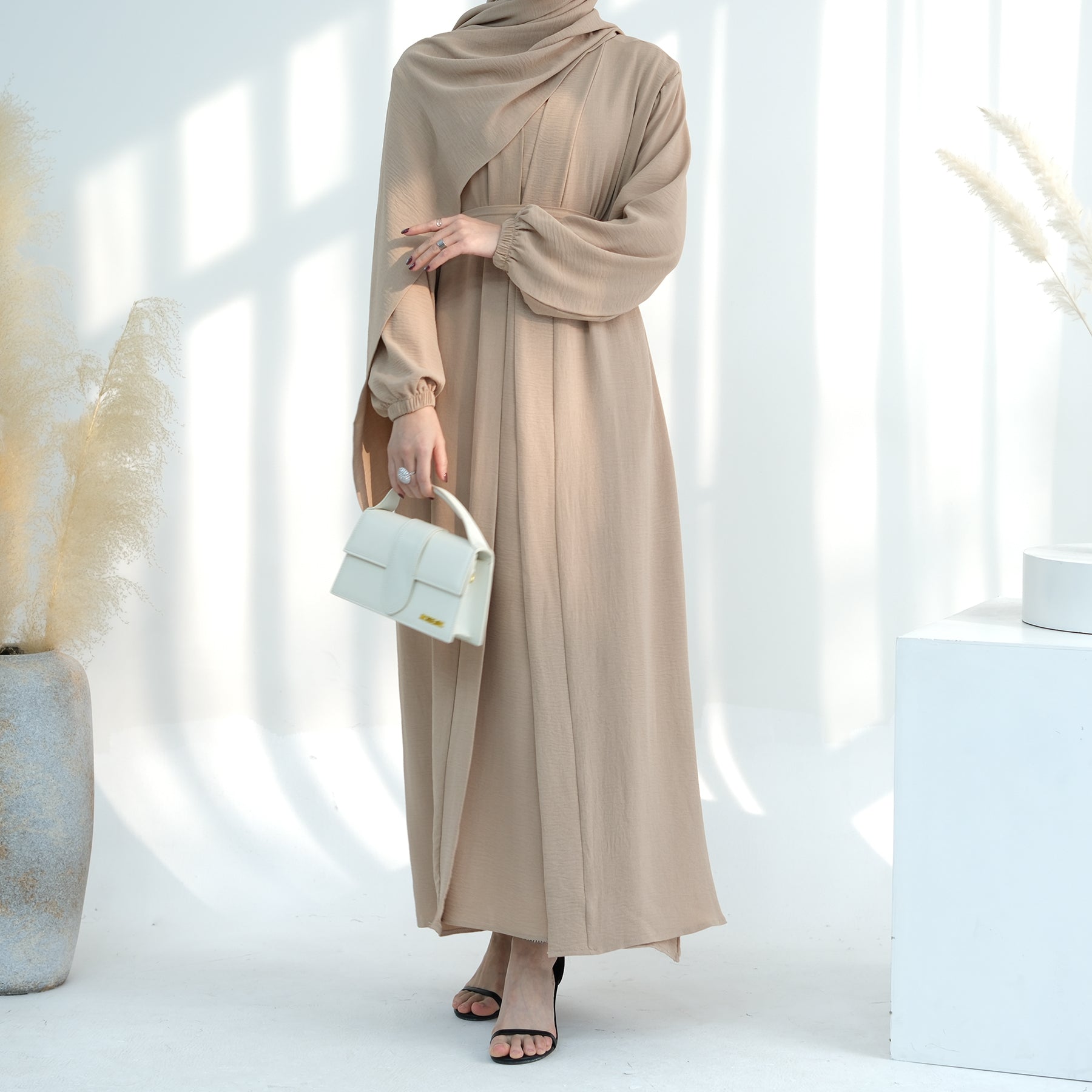 Woman wearing a beige hijab and dress holding a white handbag in a minimalistic setting. Noor Al Abaya (نور للعباية) - Noor & Grace