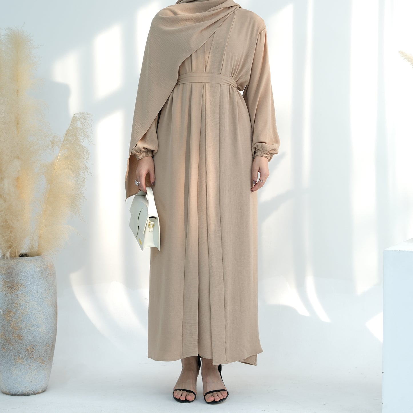 Woman wearing a beige dress and hijab standing against a white background. Noor Al Abaya (نور للعباية) - Noor & Grace