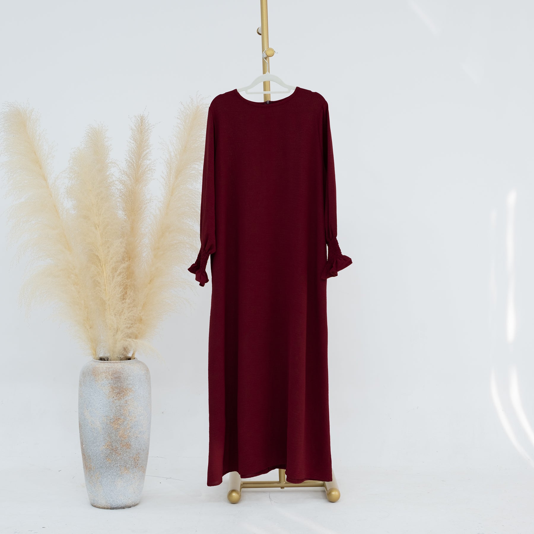 Maroon dress on a hanger with a vase of dried plants on a white background