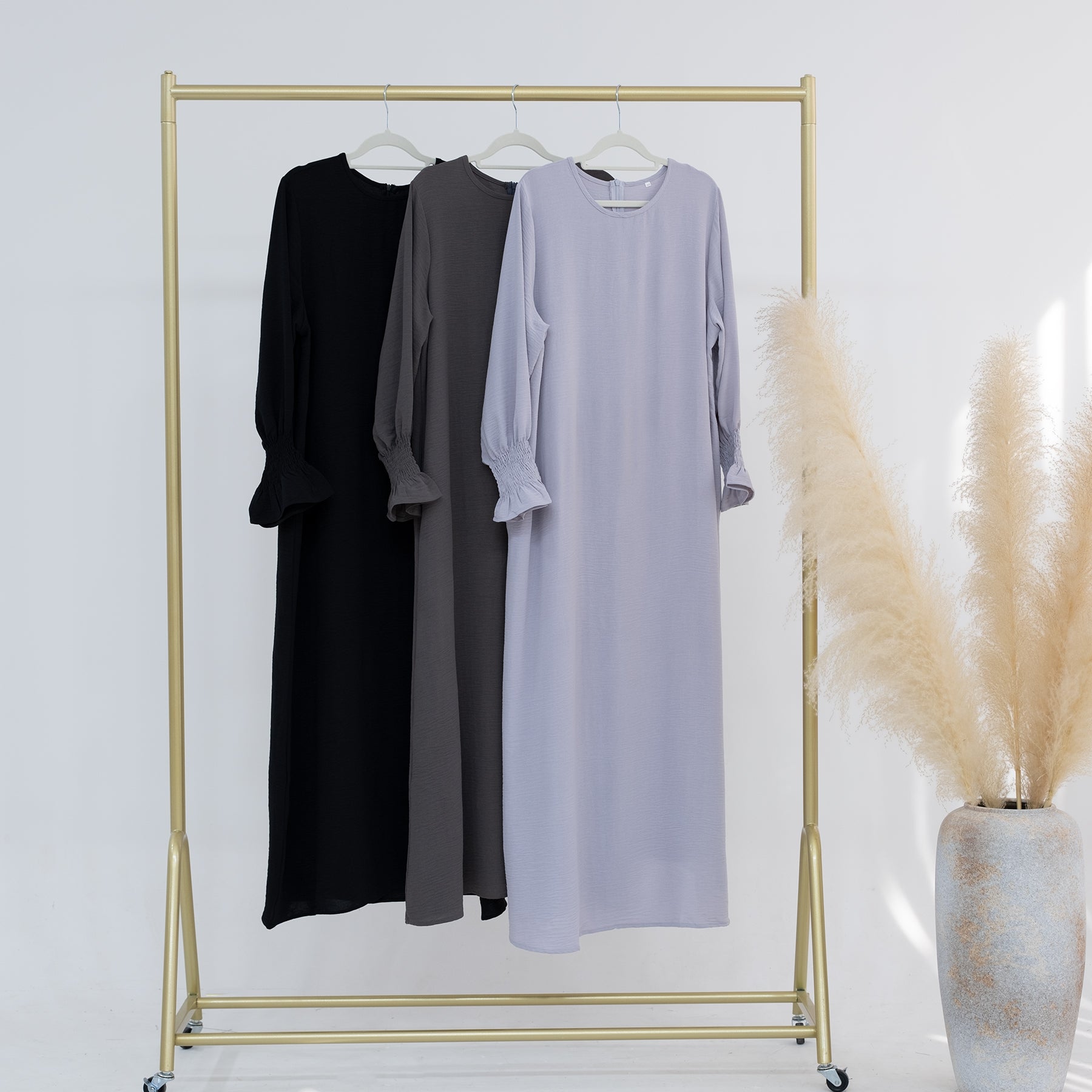 Three dresses in black, gray, and light purple hanging on a gold rack against a white wall.
