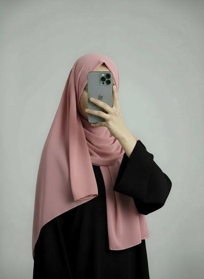 Person wearing a pink hijab and black outfit holding a phone on a gray background. Pink_Chiffon_Hijab Noor & Grace