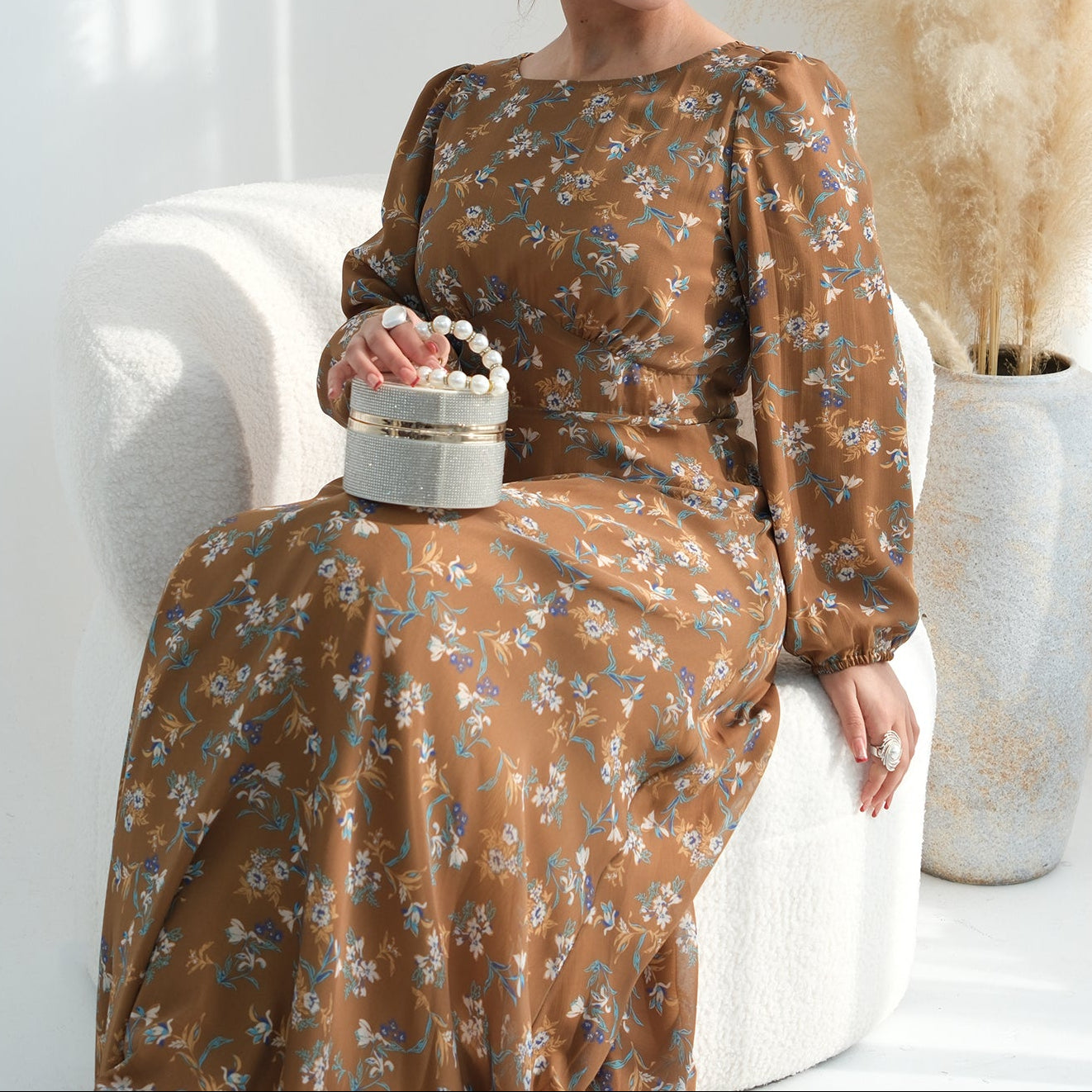 Woman in a brown floral dress sitting on a white chair with a decorative box in a bright room.