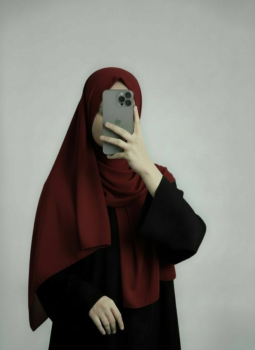 Person wearing a red hijab and black outfit holding a phone on a plain background. Red_Chiffon_Hijab Noor & Grace