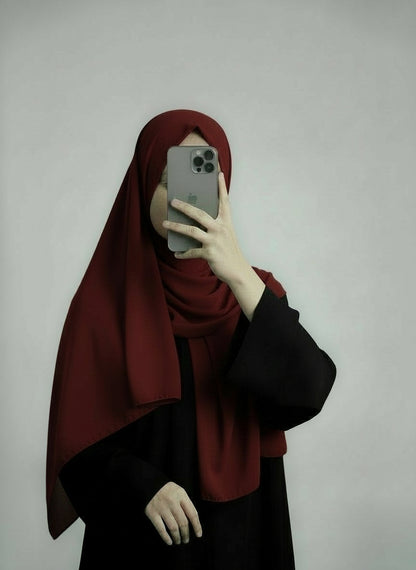 Person wearing a red hijab and black outfit holding a phone on a plain background. Red_Chiffon_Hijab Noor & Grace