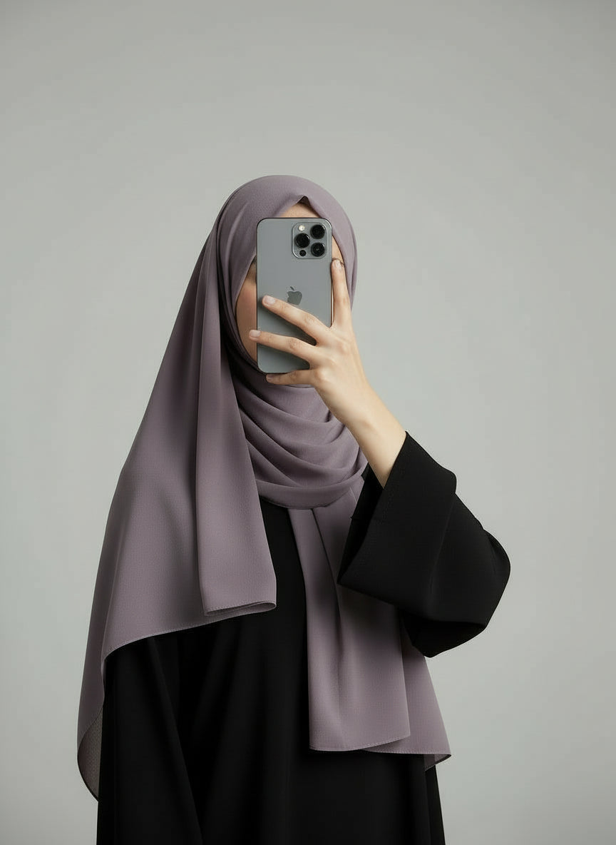 Person wearing a taro hijab and black outfit holding a phone on a gray background. Taro_Chiffon_Hijab Noor & Grace