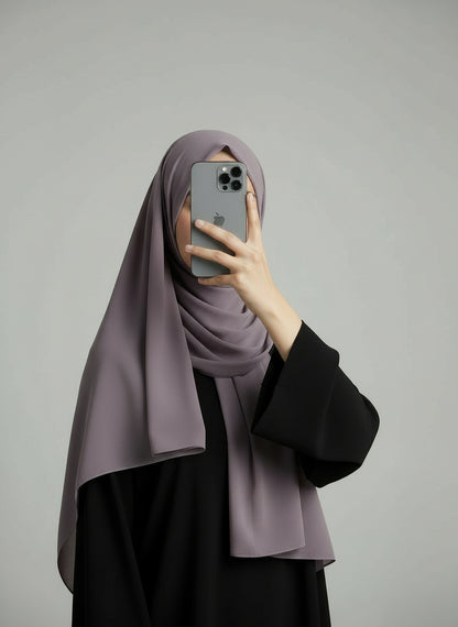 Person wearing a taro hijab and black outfit holding a phone on a gray background. Taro_Chiffon_Hijab Noor & Grace