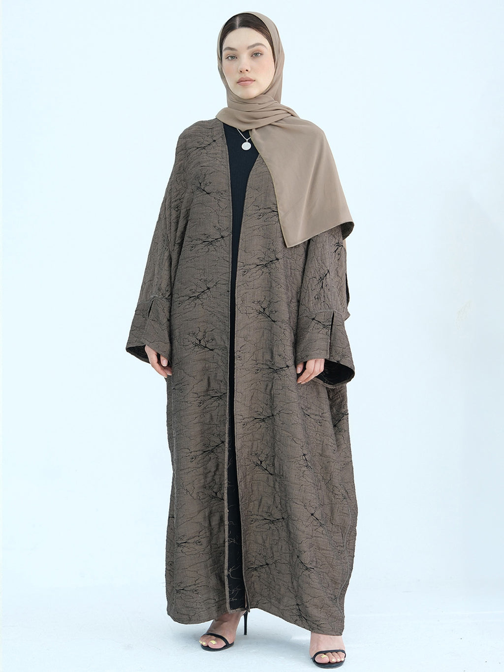 A woman modeling a long taupe-brown abaya with a subtle textured pattern, paired with a solid taupe hijab and a black inner dress, set against a plain light blue background.