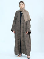 A woman modeling a long taupe-brown abaya with a subtle textured pattern, paired with a solid taupe hijab and a black inner dress, set against a plain light blue background.