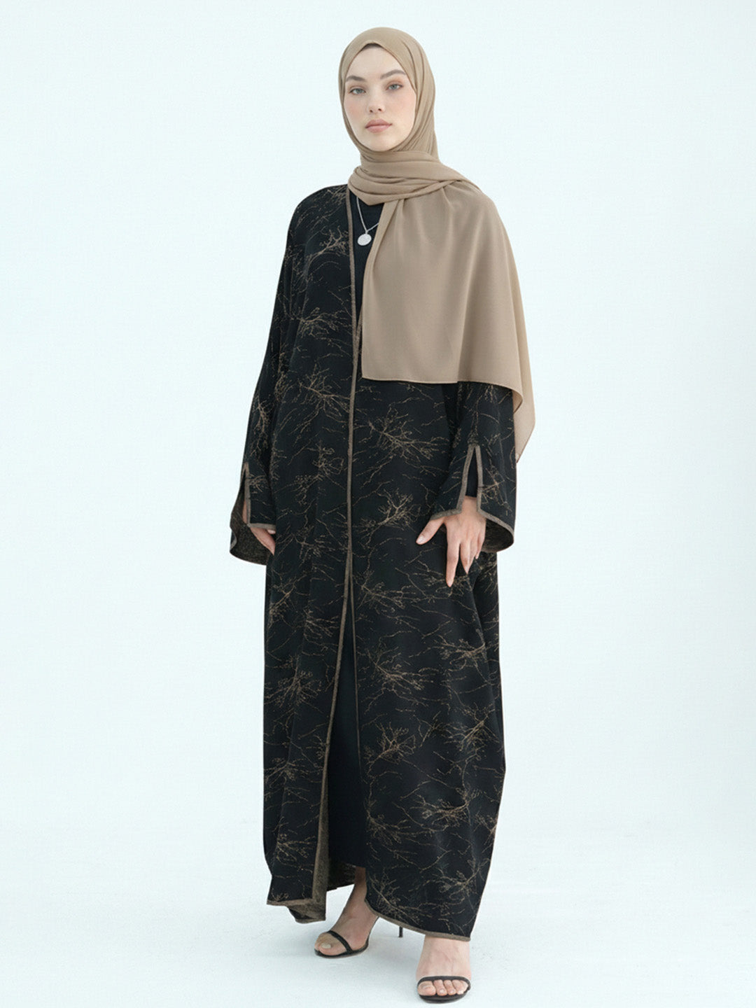 A woman modeling a long black abaya with a subtle gold abstract pattern, paired with a solid beige hijab and black heeled sandals, against a light gray studio background.