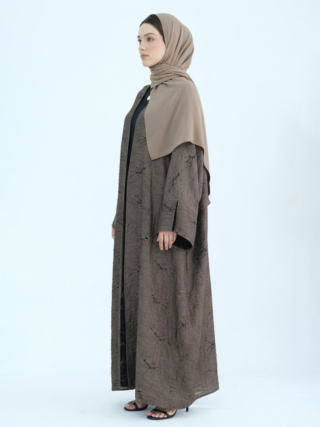 A woman wearing a long taupe-brown abaya with a subtle tonal pattern, a solid taupe hijab, and black inner lining, standing in profile against a light gray studio background.