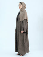 A woman wearing a long taupe-brown abaya with a subtle tonal pattern, a solid taupe hijab, and black inner lining, standing in profile against a light gray studio background.