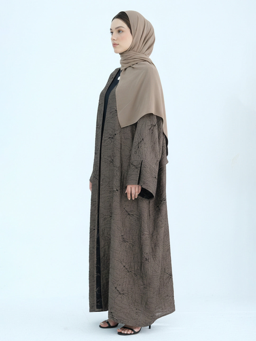 A woman wearing a long taupe-brown abaya with a subtle tonal pattern, a solid taupe hijab, and black inner lining, standing in profile against a light gray studio background.