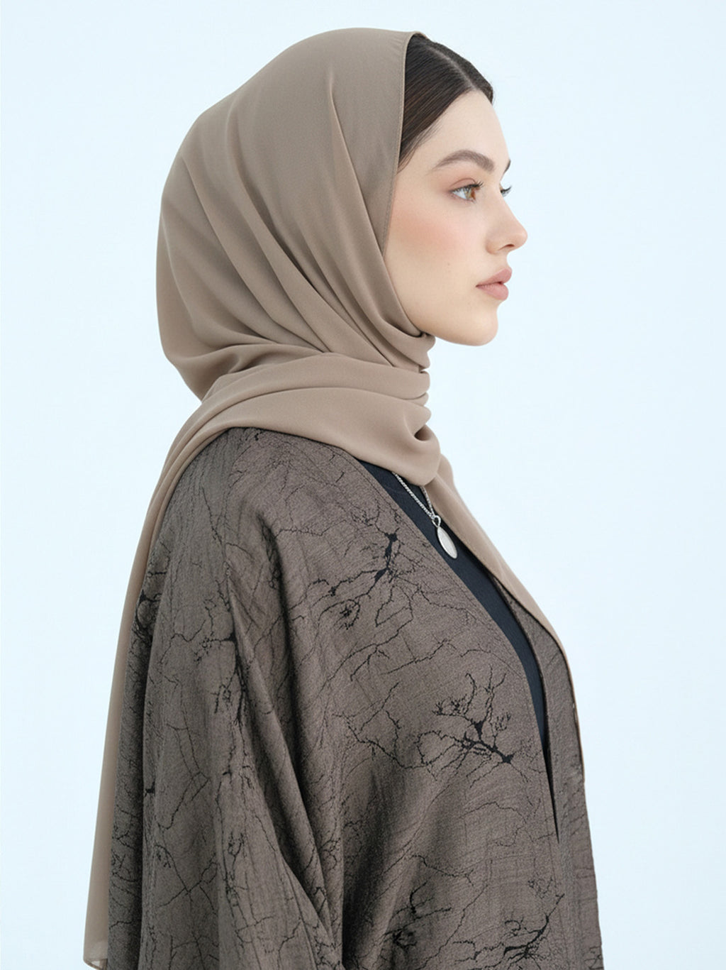 A woman in profile wearing a taupe-beige hijab and a dark brown patterned kaftan against a light blue background.