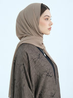 A woman in profile wearing a taupe-beige hijab and a dark brown patterned kaftan against a light blue background.