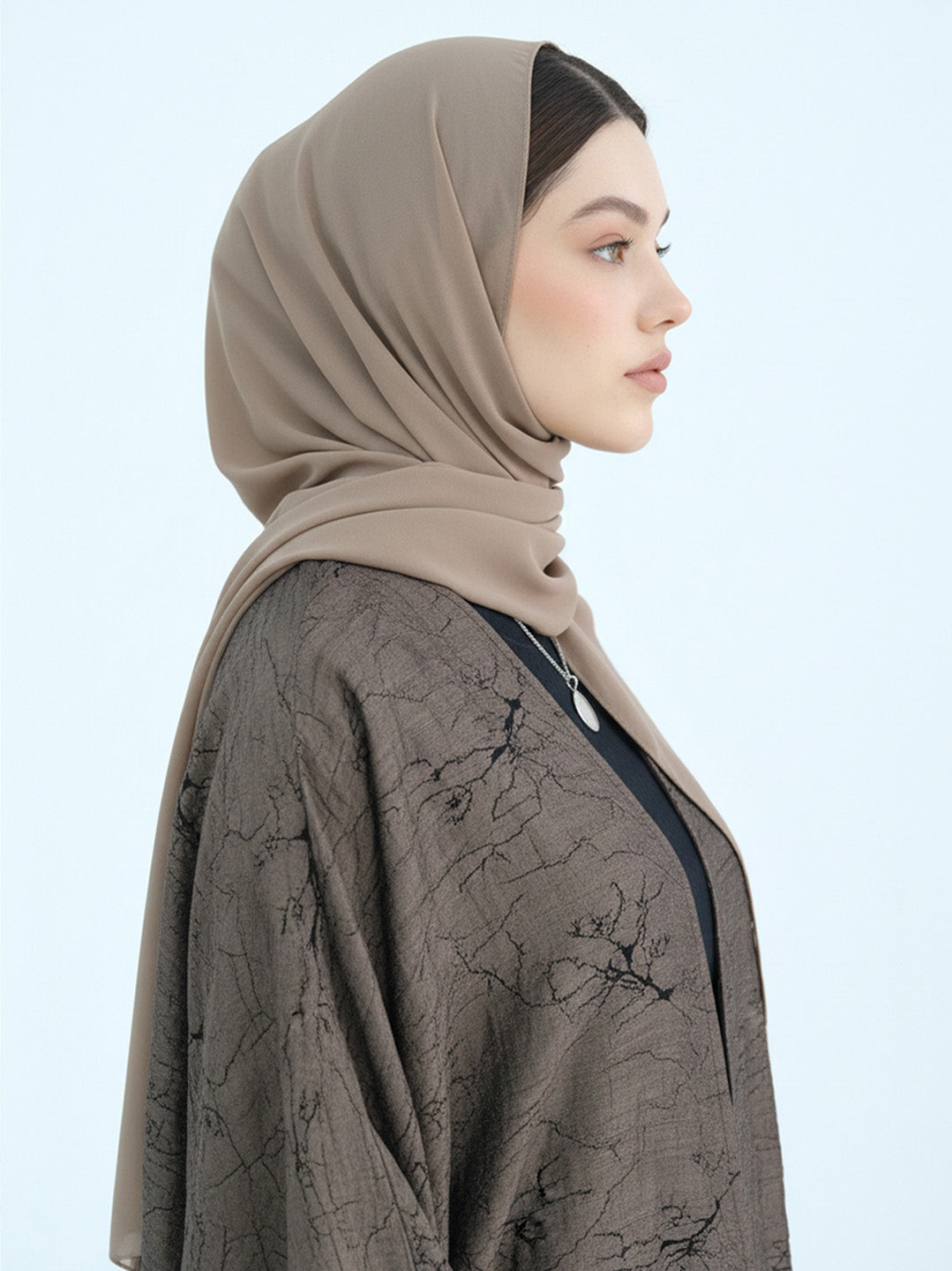 A woman in profile wearing a taupe-beige hijab and a dark brown patterned kaftan against a light blue background.