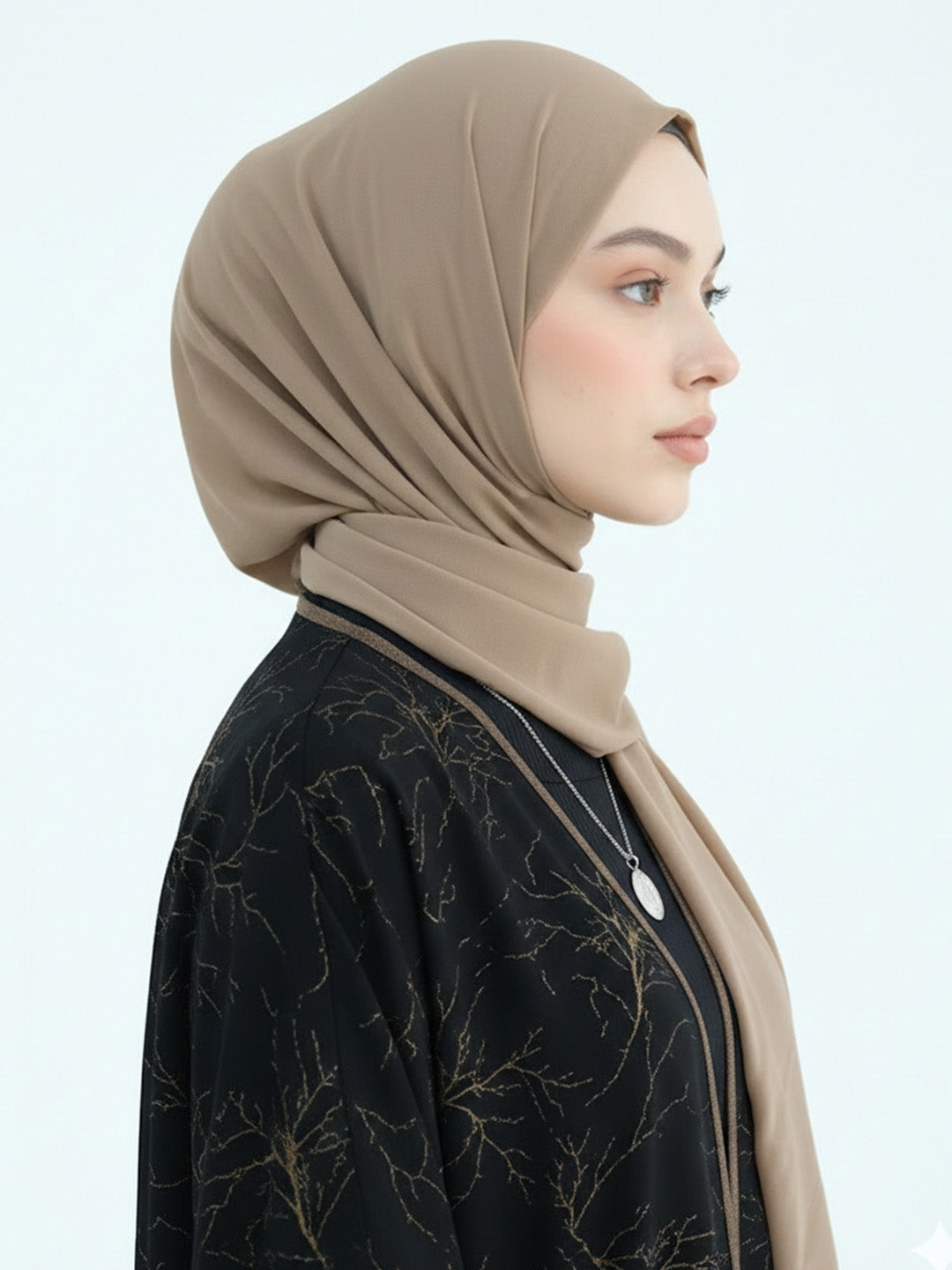 A woman in profile wearing a taupe-beige hijab and a black embroidered top, photographed against a clean light gray studio background.