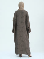 A woman viewed from the back wearing a loose, floor-length taupe-brown abaya with a subtle tonal floral pattern and a matching beige hijab, standing against a plain light gray studio background.