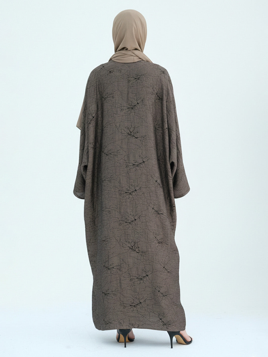 A woman viewed from the back wearing a loose, floor-length taupe-brown abaya with a subtle tonal floral pattern and a matching beige hijab, standing against a plain light gray studio background.