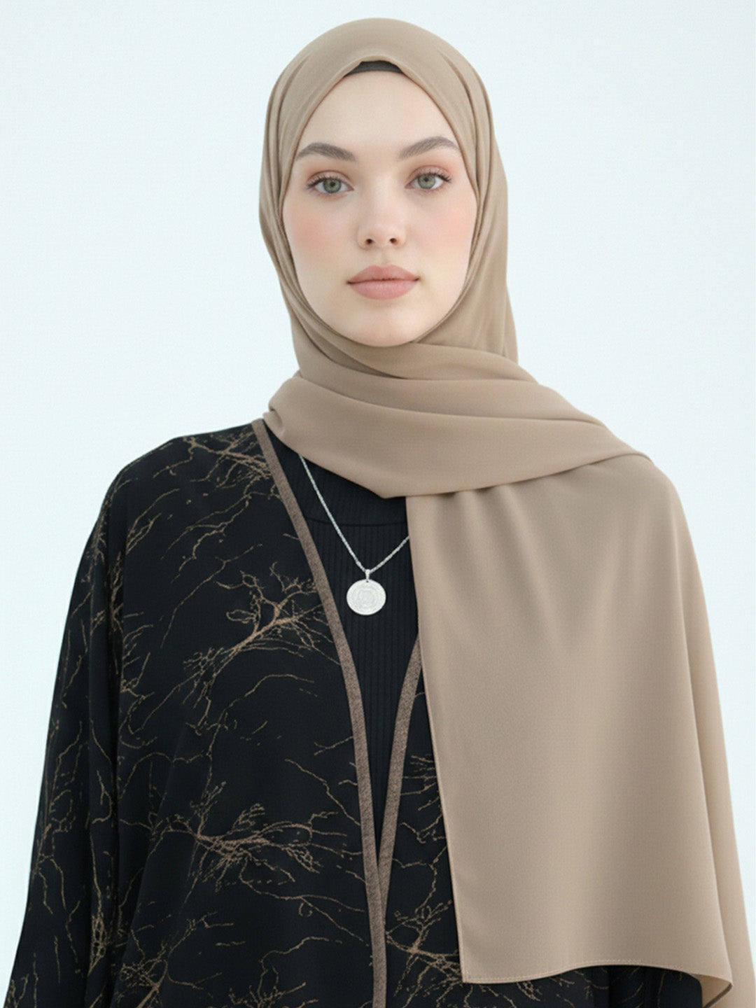 A woman wearing a taupe-beige hijab and a black long-sleeve abaya with a subtle gold branch pattern, photographed against a clean white studio background.
