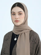 A woman wearing a taupe-beige hijab and a dark brown patterned cardigan over a black top, photographed against a light blue studio background.