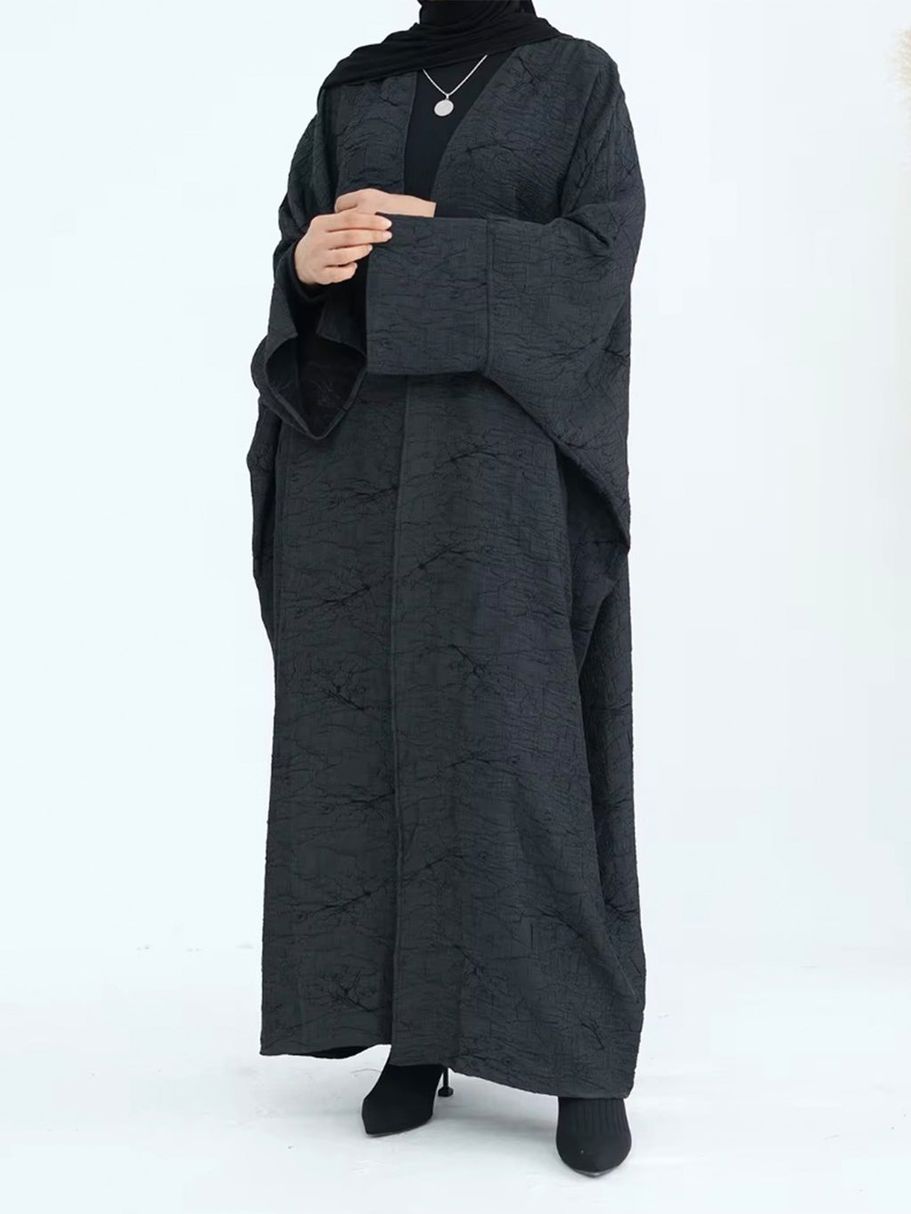 A woman wearing a long black textured abaya with wide sleeves, a black inner top, and a black headscarf, standing against a plain white studio background.