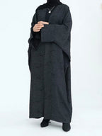 A woman wearing a long black textured abaya with wide sleeves, a black inner top, and a black headscarf, standing against a plain white studio background.