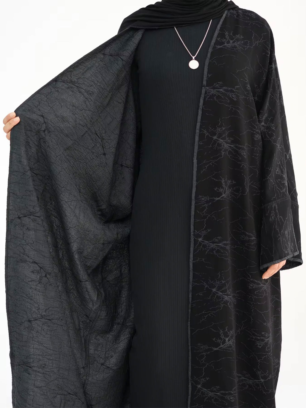 A black abaya dress with a subtle marble-like textured pattern, worn by a model and styled with a matching black hijab and a silver pendant necklace.