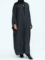 A person wearing a long, loose black abaya with a subtle textured pattern, a black turtleneck underneath, a black hijab, and black pointed-toe heels against a plain white studio background.
