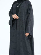 A woman wearing a dark charcoal gray textured abaya with a black inner top and a black headscarf, shown from the neck down against a plain white background.