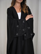 A woman wearing a black abaya with wide sleeves, featuring a contrasting inner lining with embroidered silver bow motifs and small rhinestone accents, styled over a white top.