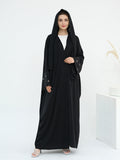 A woman modeling a long black abaya with a matching black hood and sheer embroidered sleeves, worn over a white top, in a minimalist white studio setting.