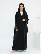 A woman modeling a long black abaya with a matching black hood and sheer embroidered sleeves, worn over a white top, in a minimalist white studio setting.