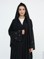 A woman wearing a black abaya with a matching black head covering and long sleeves, featuring delicate silver embroidery on the cuffs, over a white inner top. She is standing against a plain white studio background.