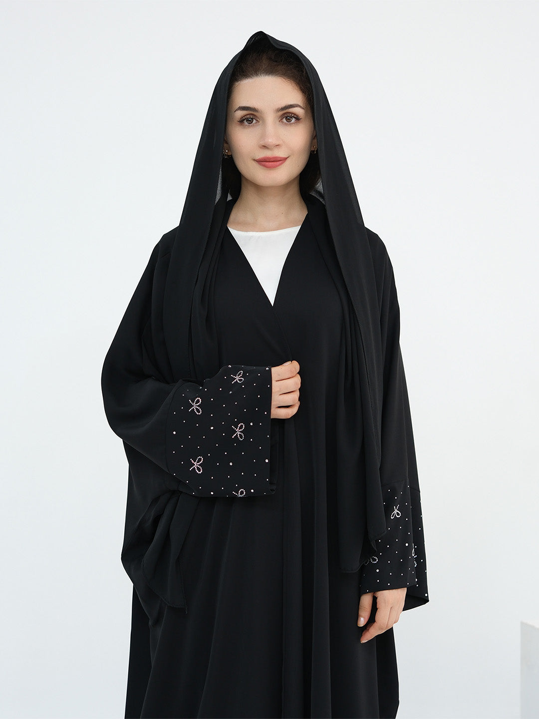 A woman wearing a black abaya with a matching black head covering and long sleeves, featuring delicate silver embroidery on the cuffs, over a white inner top. She is standing against a plain white studio background.