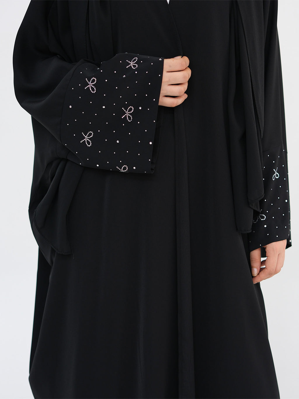 A close-up of a black abaya dress featuring wide sleeves with decorative cuffs embellished with small white dots and bow motifs, shown on a model against a light gray background.