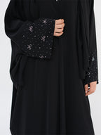 A close-up of a black abaya dress featuring wide sleeves with decorative cuffs embellished with small white dots and bow motifs, shown on a model against a light gray background.