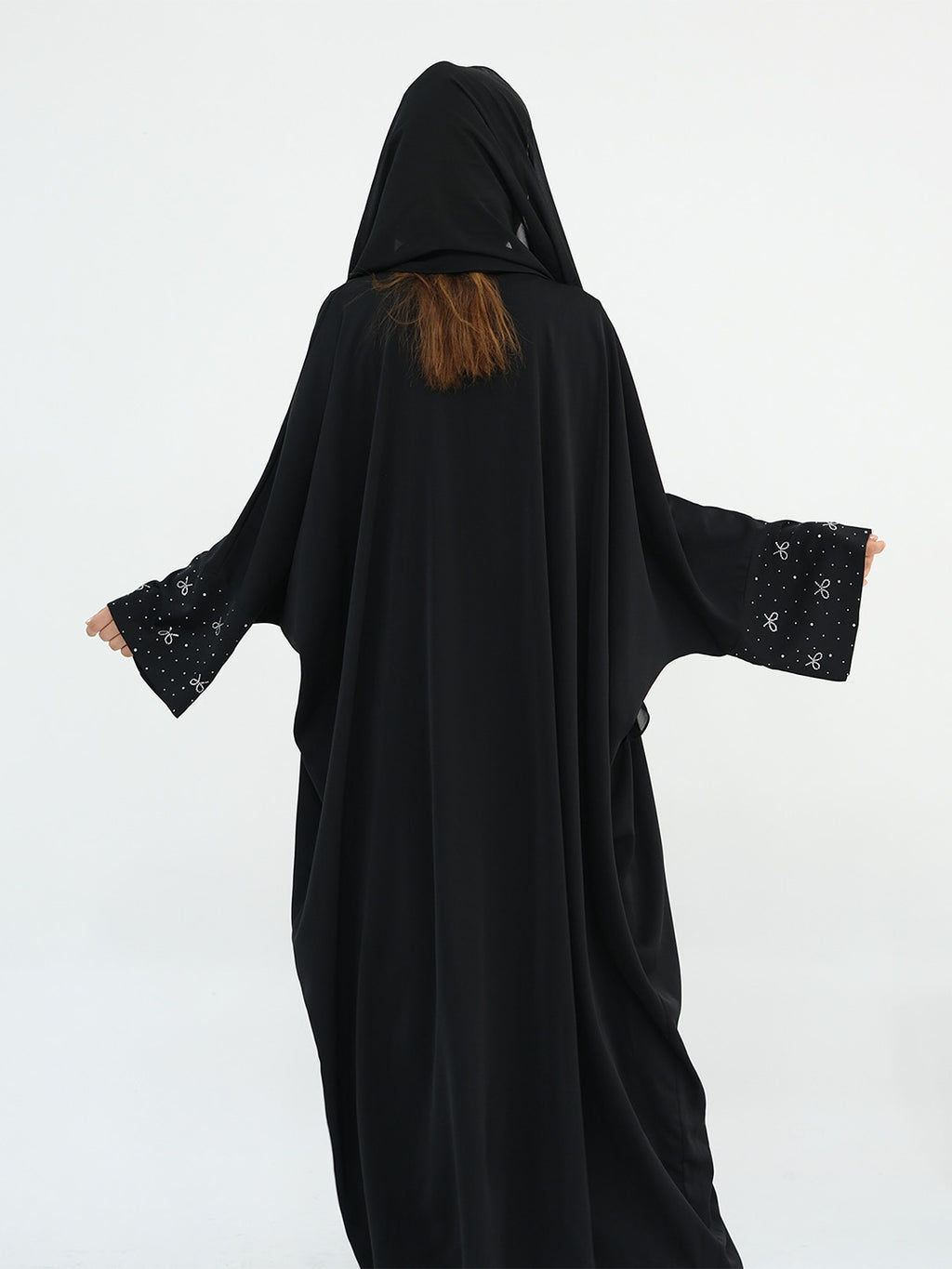 A person wearing a black hooded abaya or long robe with wide sleeves, shown from the back against a plain light gray studio background. The sleeves have small light embellishments near the cuffs, and a small section of brown hair is visible at the neckline.