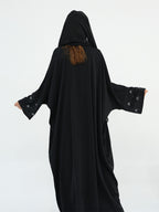 A person wearing a black hooded abaya or long robe with wide sleeves, shown from the back against a plain light gray studio background. The sleeves have small light embellishments near the cuffs, and a small section of brown hair is visible at the neckline.