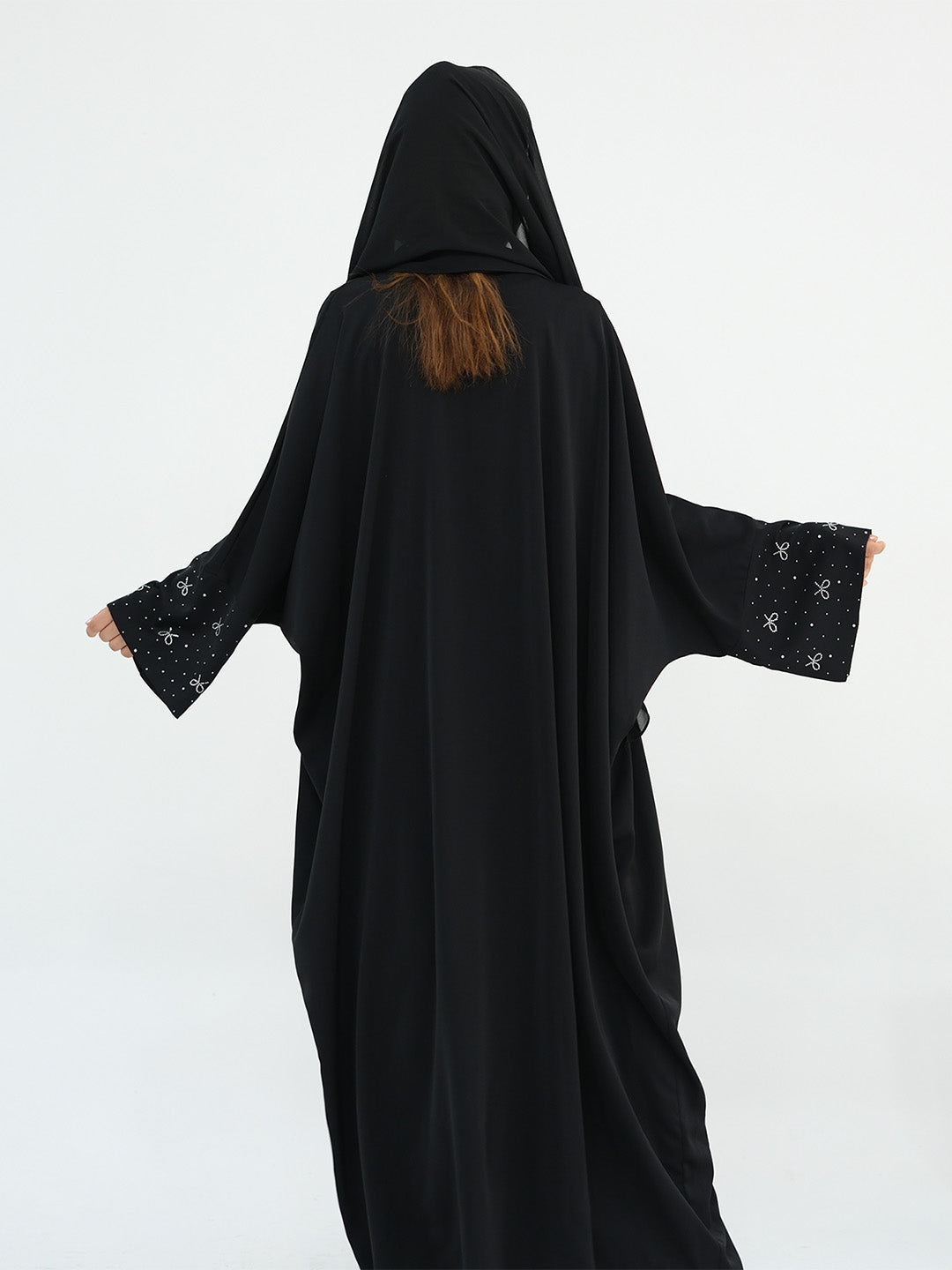 A person wearing a black hooded abaya or long robe with wide sleeves, shown from the back against a plain light gray studio background. The sleeves have small light embellishments near the cuffs, and a small section of brown hair is visible at the neckline.
