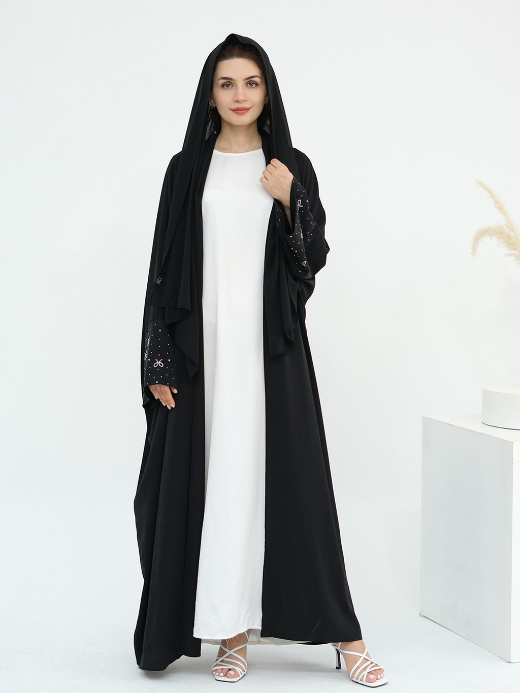 A woman modeling a long black abaya with sheer embroidered sleeves over a white maxi dress, standing in a bright white studio setting.