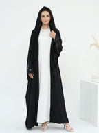 A woman modeling a long black abaya with sheer embroidered sleeves over a white maxi dress, standing in a bright white studio setting.