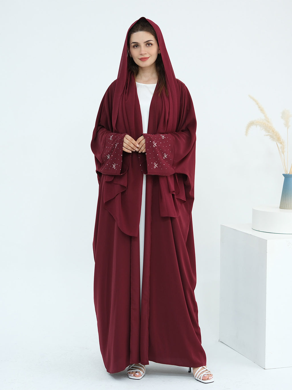 A woman modeling a deep burgundy hooded abaya with embroidered details on the sleeves, worn over a white inner top. She is standing in a bright, minimalist studio setting with a white pedestal and dried pampas grass in a vase.