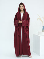 A woman modeling a deep burgundy hooded abaya with embroidered details on the sleeves, worn over a white inner top. She is standing in a bright, minimalist studio setting with a white pedestal and dried pampas grass in a vase.
