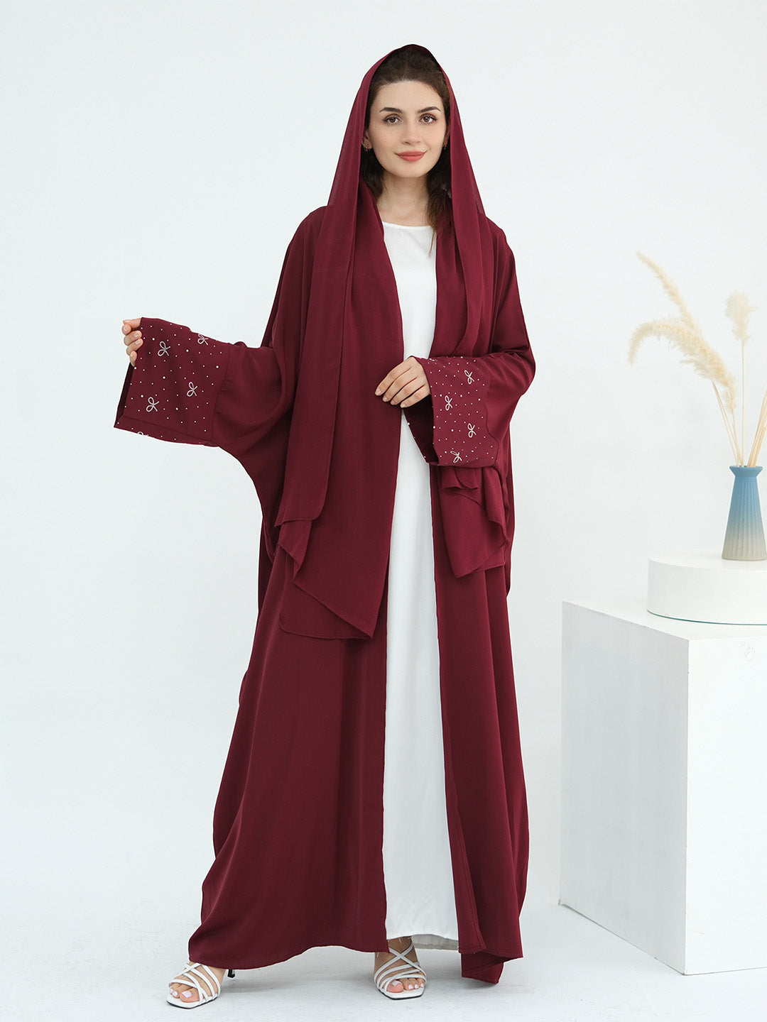 A woman models a long maroon hooded abaya with embroidered details on the sleeves, worn over a white dress, in a bright studio setting.